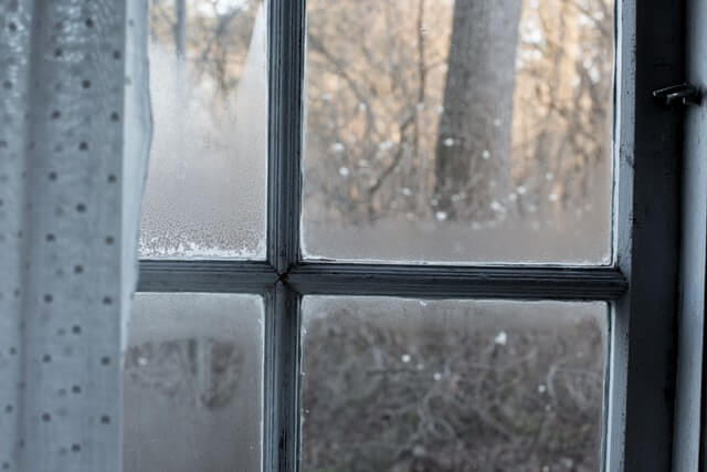 Water condenses on a wooden window that has a loose drape on the interior.